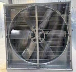 A stainless steel greenhouse exhaust fan mounted on the wall of a glass greenhouse, actively ventilating air to regulate temperature. The fan features automatic shutters and is surrounded by lush green plants thriving in a controlled environment.