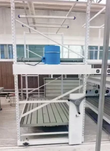 A multi-tiered metal rolling rack holding numerous trays of young, green seedlings. The rack is on caster wheels, highlighting its mobility. Each shelf is filled with organized propagation trays, showcasing a space-efficient system for starting seeds in a greenhouse or nursery.